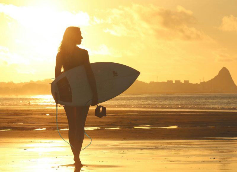 A silhouette of a woman holding a surfboard, standing on a beach at sunset with a golden sky.