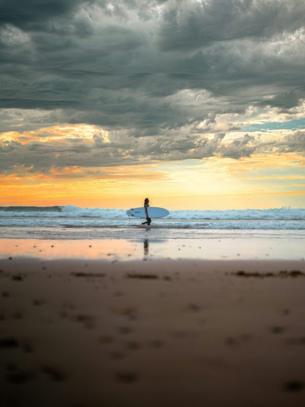 A serene view of a surfer walking along the beach with a surfboard at sunset in Essaouira, Morocco.