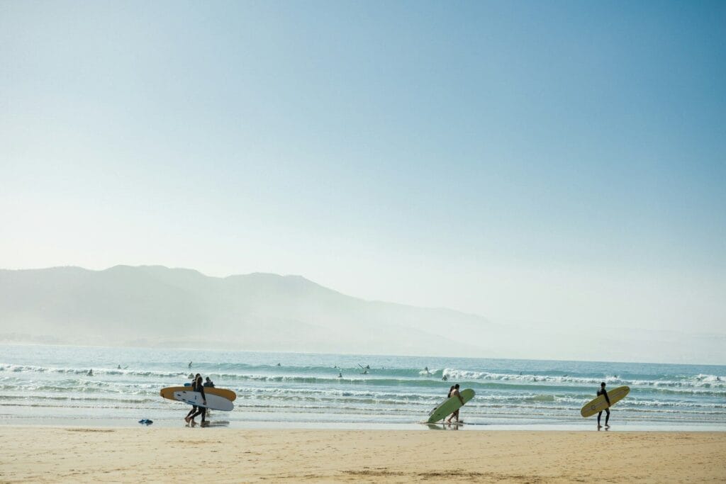 a group of people carrying surfboards on top of a beach.August Weather in Morocco